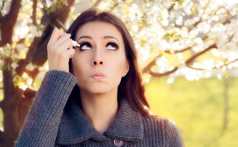 Young woman with allergies and itchy eyes stands outside while putting eye drops in her eyes.